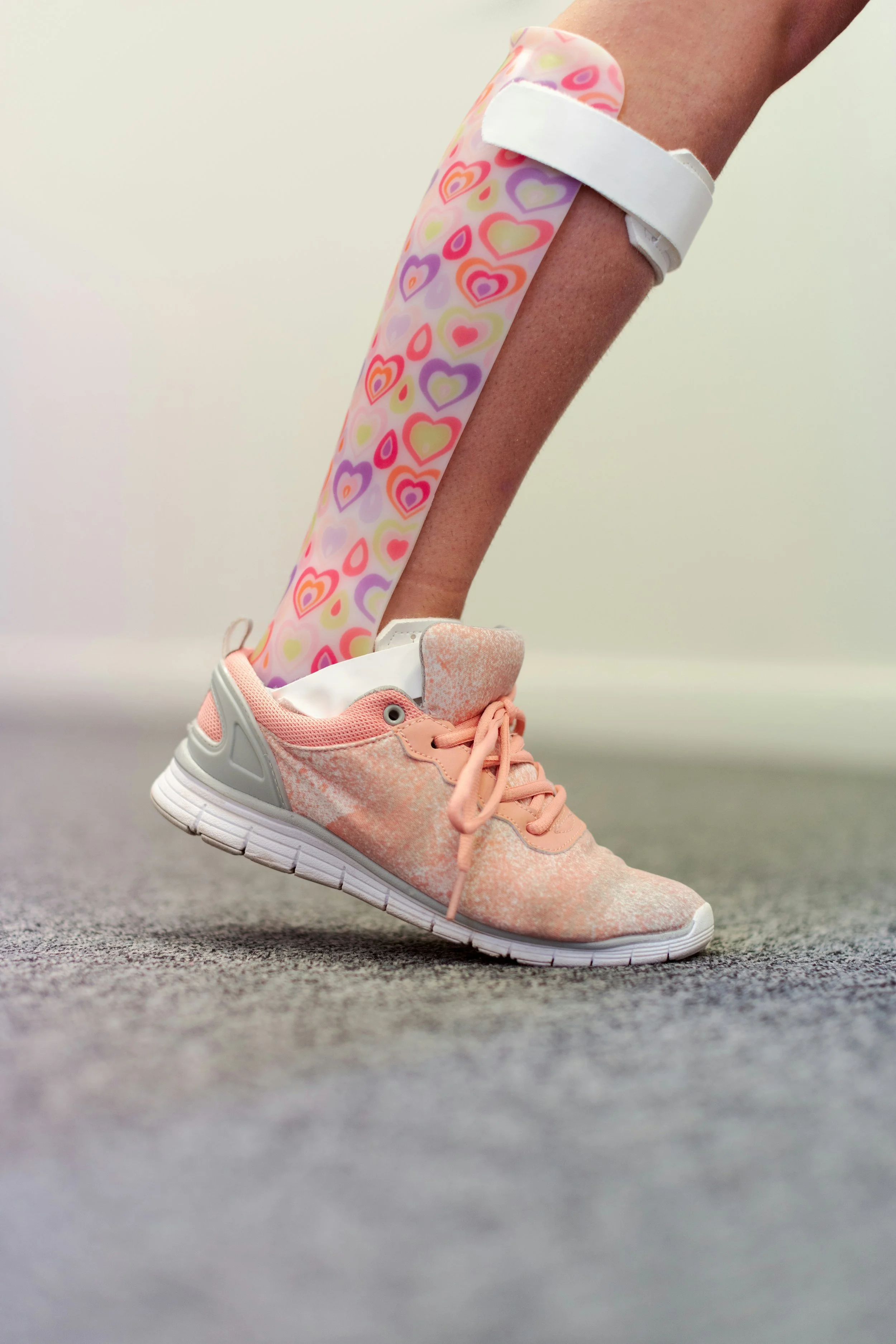 A person wearing pink athletic shoes, standing on a gray carpeted floor, with their leg extended and a heart-patterned bandage on their lower calf.