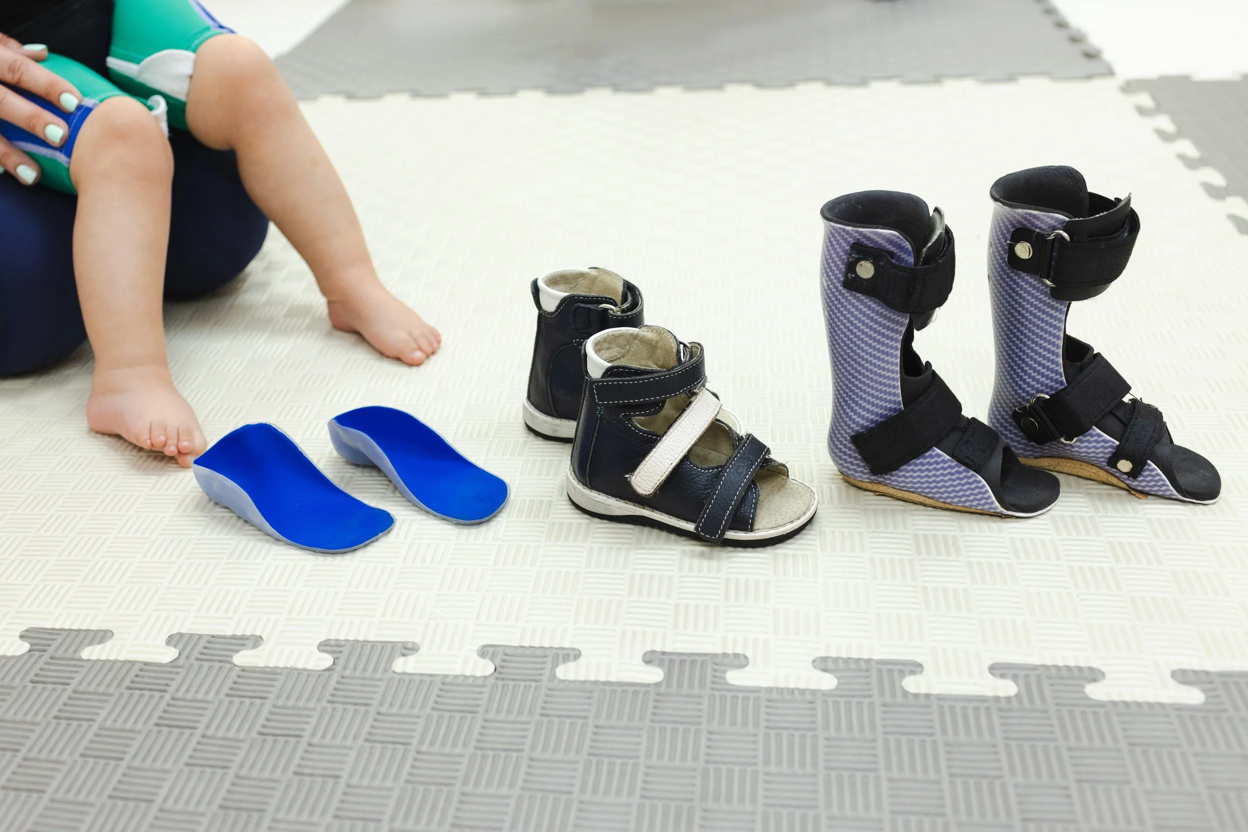 Child sitting on the floor with legs crossed, surrounded by various pairs of shoes including black sandals, gray patterned boots, and blue slippers, on a padded mat.