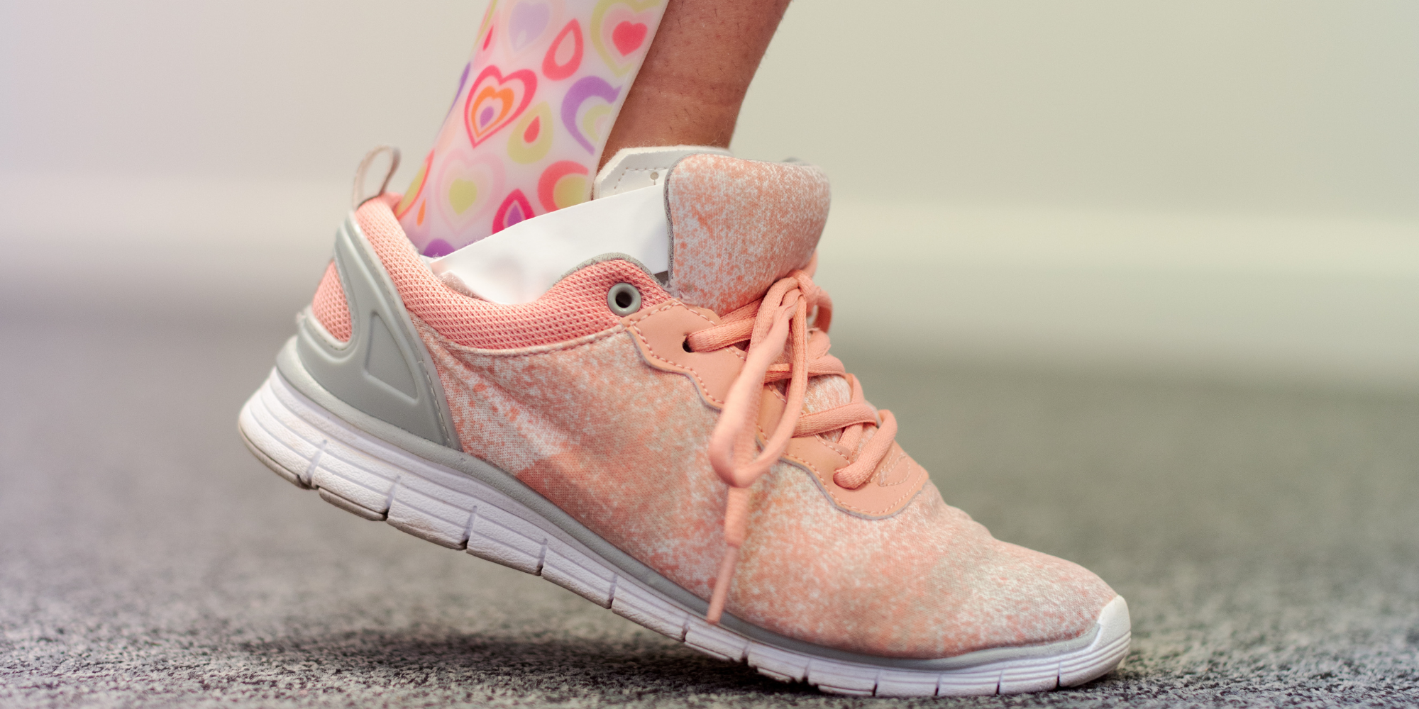 Close-up of a person wearing a peach-colored athletic shoe with pink laces, white sole, and gray accents, standing on a textured surface, with a colorful sock featuring heart patterns visible above the shoe.