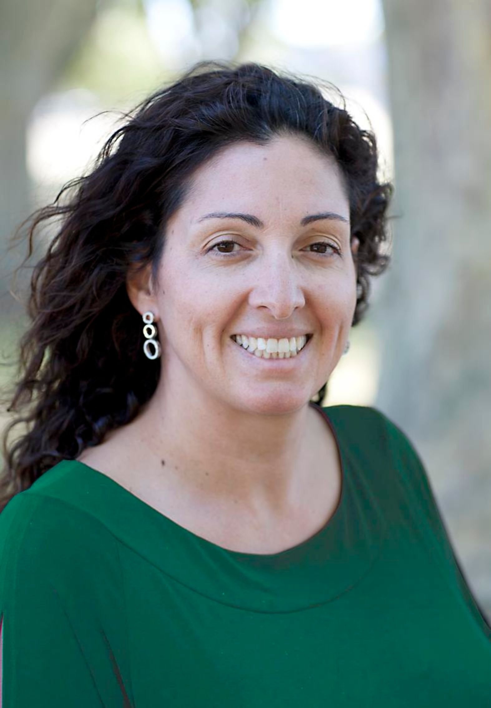 Close-up of a smiling woman with curly dark hair, wearing earrings and a green top, outdoors with blurred background.