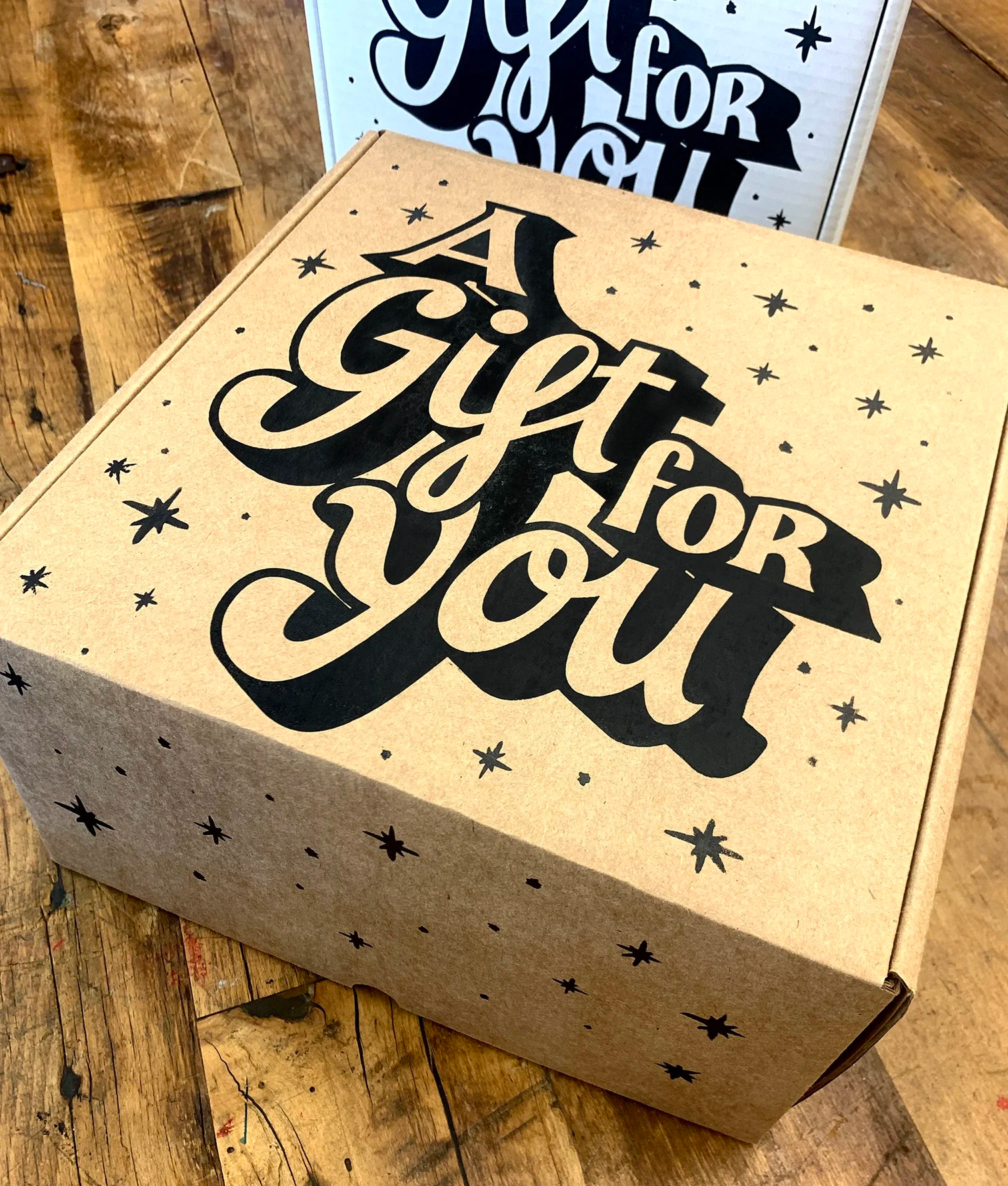 A cardboard gift box with the phrase 'A Gift for You', screenprinted in large black script on the lid, surrounded by small black star illustrations
