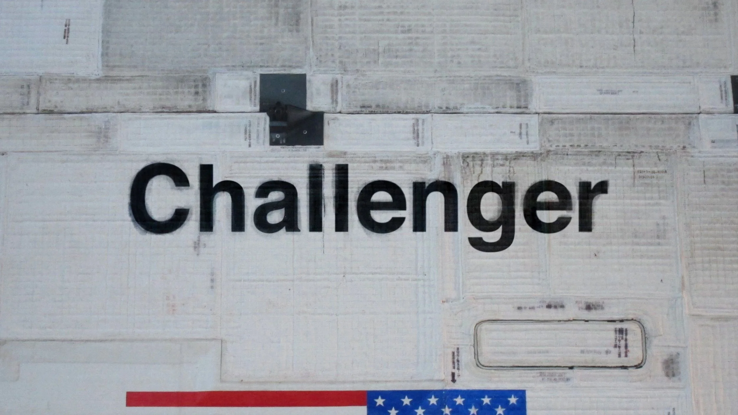 Close-up image of the word "Challenger" on a textured surface resembling wood or paper, with a partial American flag at the bottom.