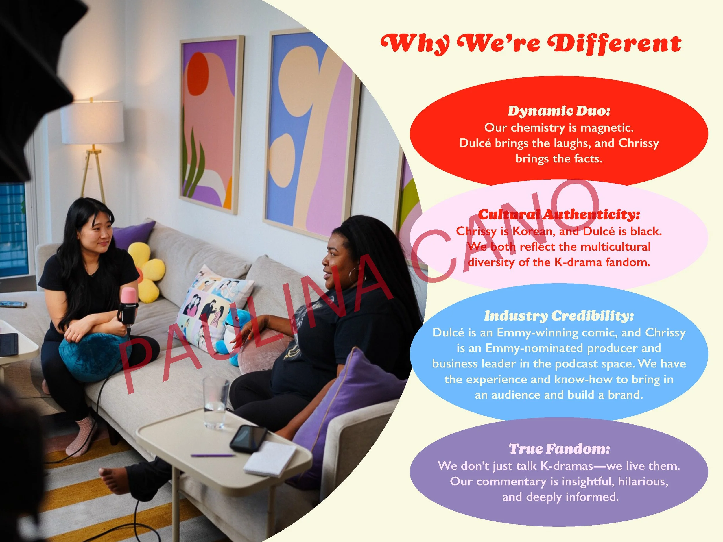 Two women sitting on a beige sofa in a modern living room during an interview or podcast, with colorful abstract artwork on the wall behind them. One woman has long black hair, is holding a microphone, and wears a black shirt and blue jeans. The othe