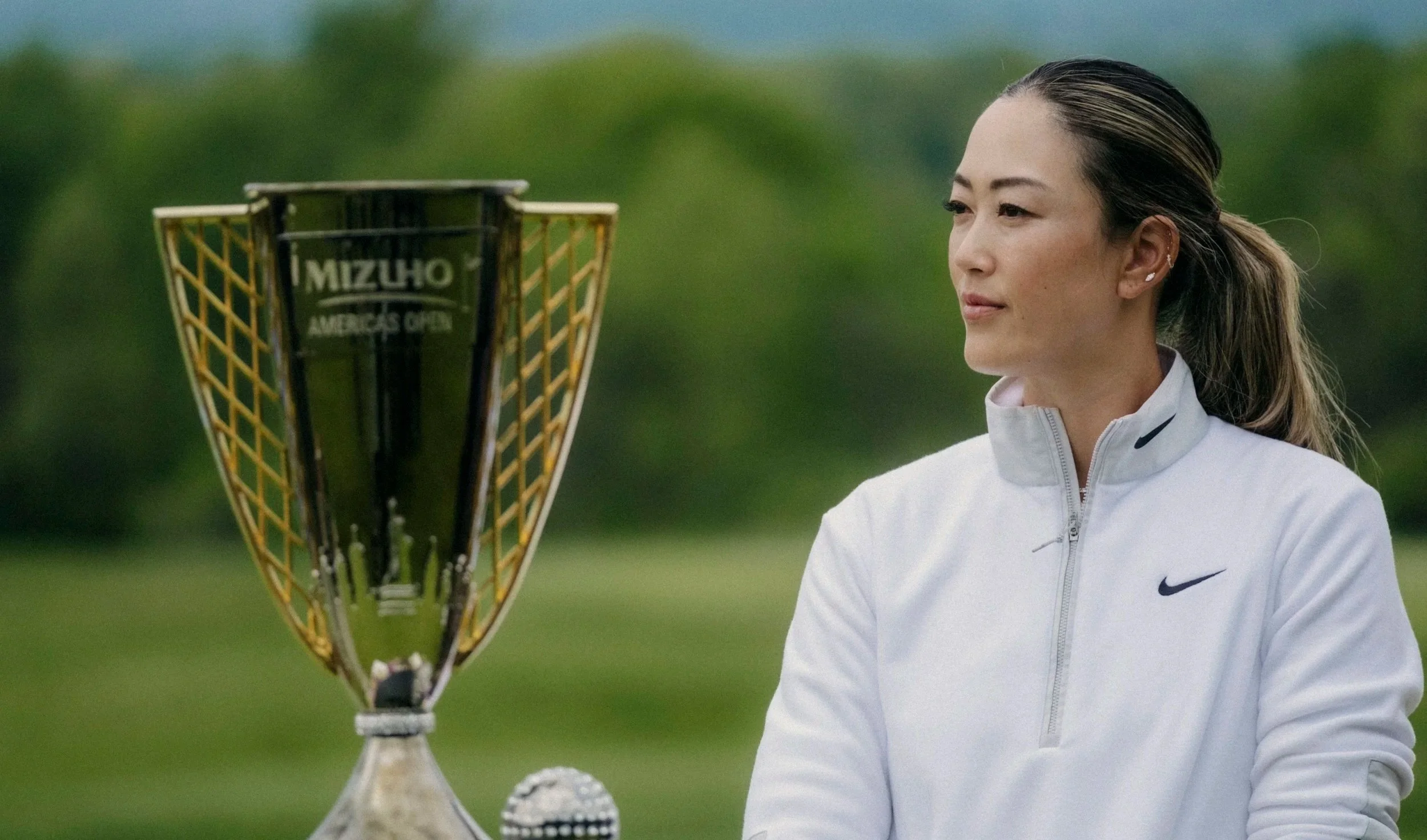 A female golfer in a white Nike zip-up jacket stands outdoors with a large golf trophy and golf ball in front of her, on a lush green background.