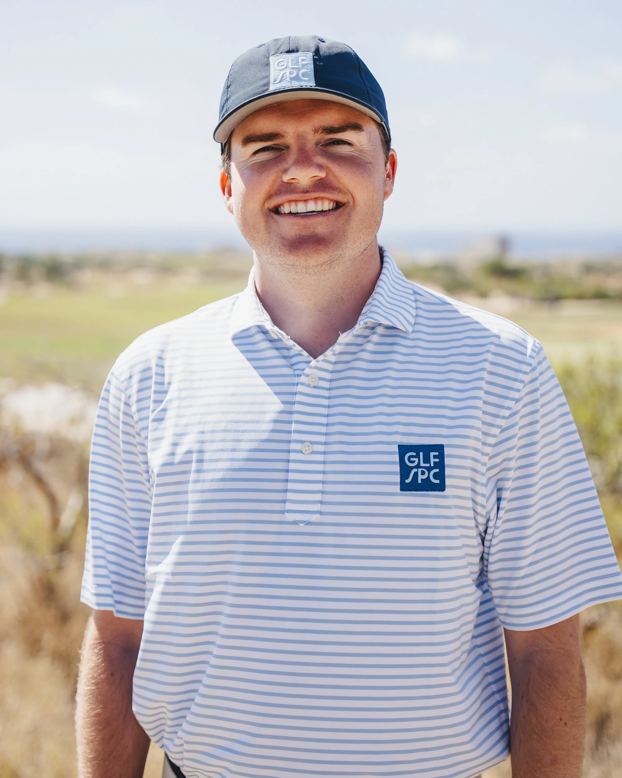 Close-up of a smiling man wearing a blue striped golf shirt and a blue cap with a logo, outdoors on a sunny day.