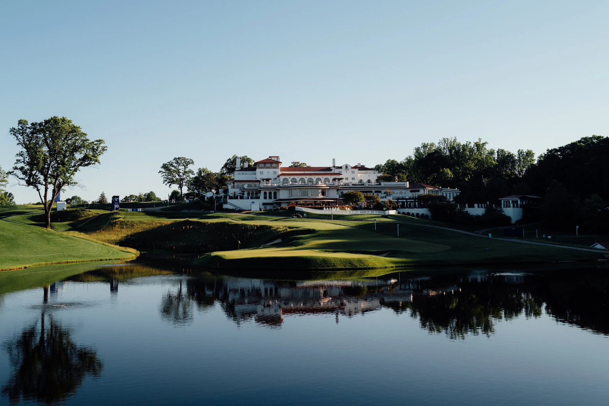 A large white building with multiple levels and red-tiled roofs is situated on a hill overlooking a golf course. The golf course features green fairways, trees, and a water hazard, with reflections visible in the calm water.
