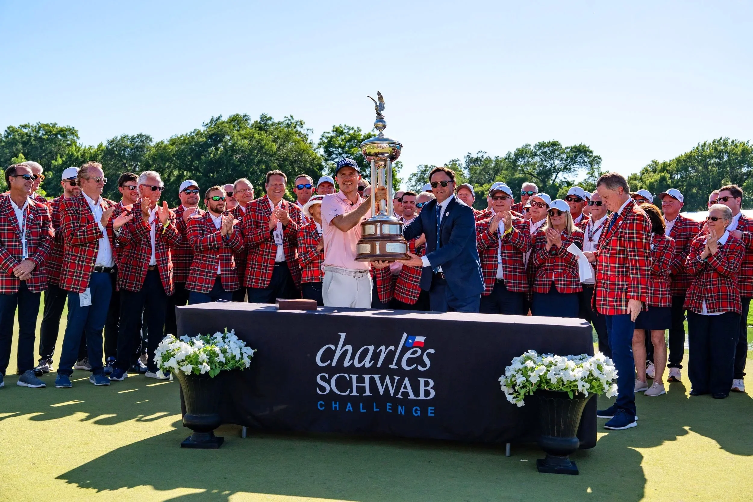 A group of men and women in red plaid jackets and white caps celebrate on a golf course. A man in a pink shirt and baseball cap holds a trophy while another man in a suit and sunglasses presents it. The backdrop shows trees and a bright blue sky, and a table in front displays a sign with the name 'Charles Schwab Challenge' decorated with white flowers.