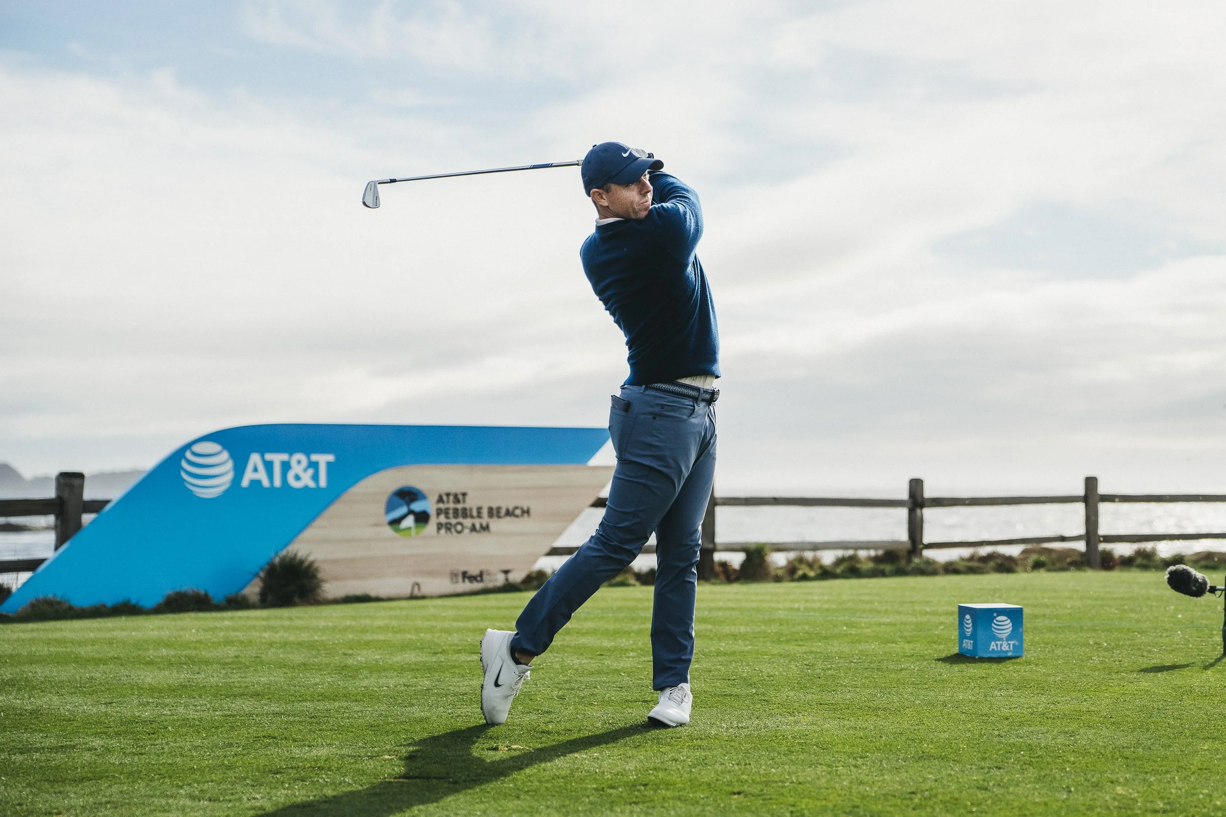A male golfer in a blue sweater and hat swings a golf club on a golf course with green grass, with AT&T and Pebble Beach signage in the background.
