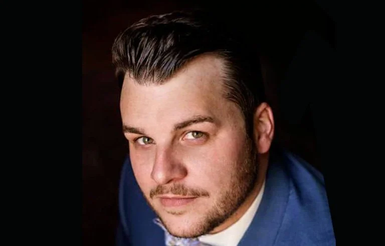 A young man with dark hair styled back, light eyes, and a light beard, wearing a blue suit and white shirt, looking at the camera.