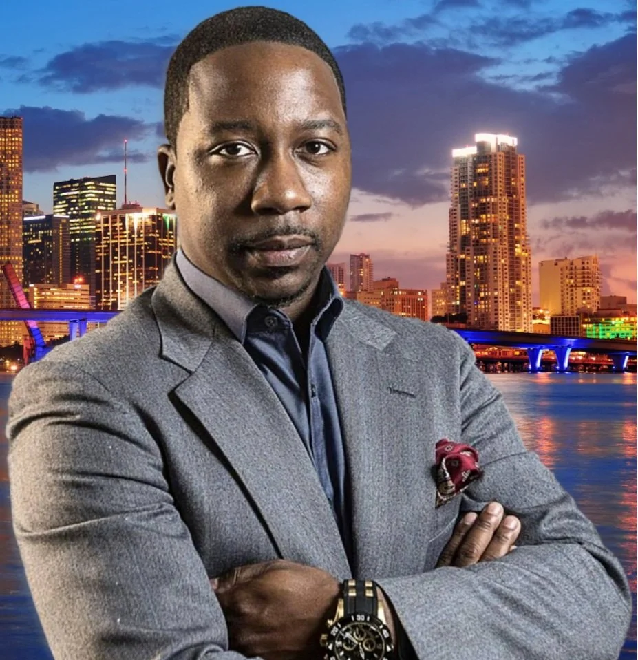 A man in a gray blazer and shirt posing with arms crossed in front of a city skyline at dusk, with buildings and a bridge reflected in the water.
