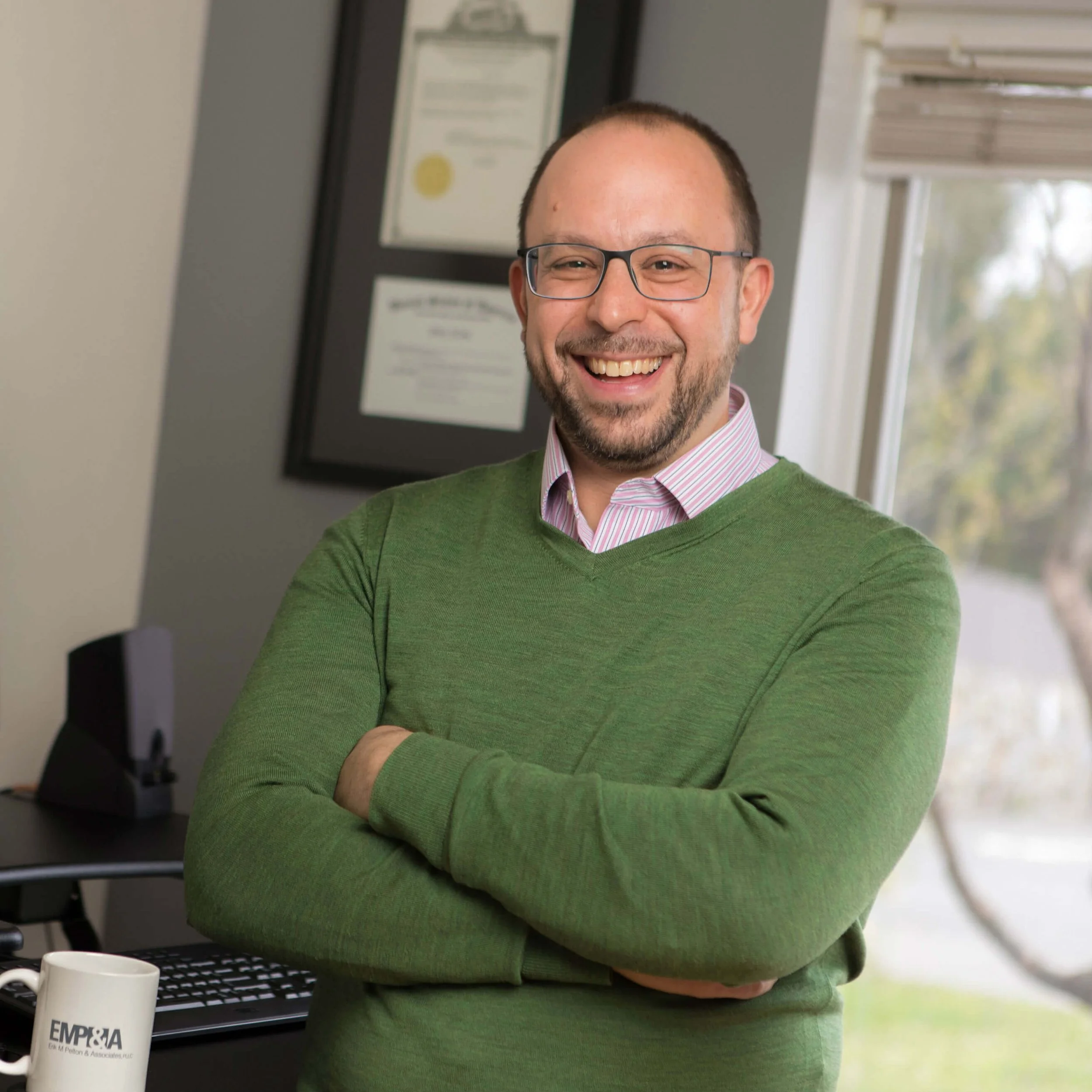 A smiling man with glasses, wearing a green sweater over a striped collared shirt, standing with crossed arms in an office space near a window, with framed certificates on the wall behind him.