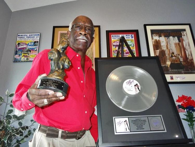 A man in a red shirt holding a music award trophy, standing next to a framed platinum record plaque on display, with framed posters and awards hanging on the wall behind him.