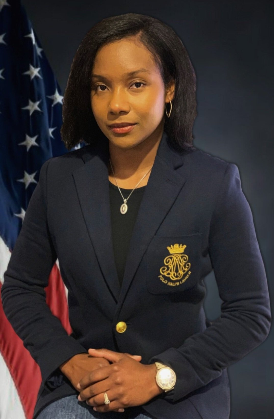 A woman wearing a navy blazer with an embroidered coat of arms, gold lapel pin, and jewelry, sitting with her hands clasped, with American flags in the background.