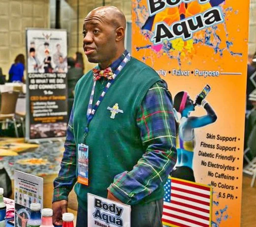 A man in a green vest with a plaid shirt and bow tie standing in front of a booth labeled 'Body Aqua' at a trade show or expo.