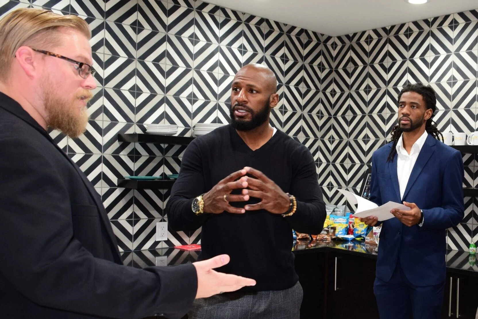 Three men in a discussion in an office break room with black and white geometric wallpaper. One man with a beard, wearing glasses and a suit, is talking to a bald man wearing a black sweater, who is making a gesture with his hands. The third man with dreadlocks, dressed in a blue suit and holding papers, listens attentively.