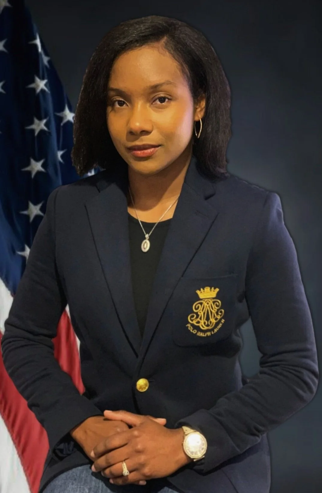 A woman wearing a navy blazer with an embroidered coat of arms, gold lapel pin, and jewelry, sitting with her hands clasped, with American flags in the background.
