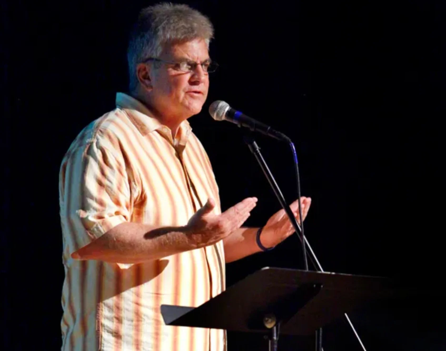 A man with gray hair and glasses stands behind a music stand, speaking into a microphone while using hand gestures, against a dark background.