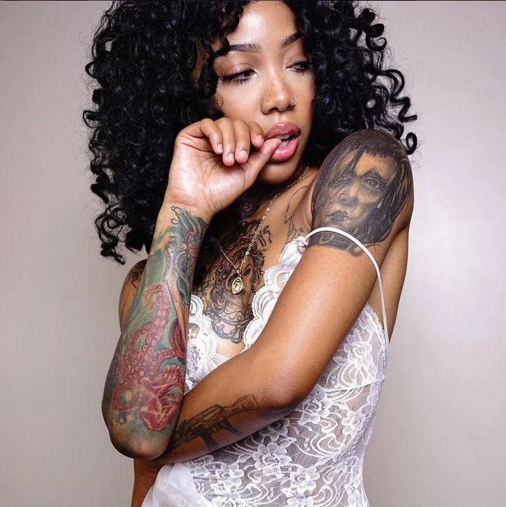 Woman with curly black hair and multiple tattoos, wearing a white lace top, posing with her hand near her mouth against a neutral background.