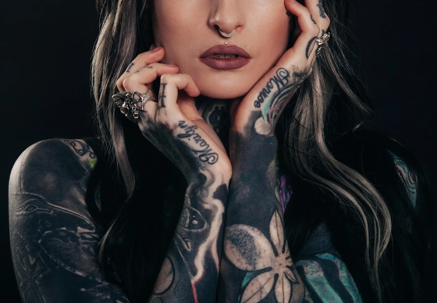 Close-up of a person with tattoos and piercings, displaying hands with rings, showcasing extensive body art, and wearing makeup.