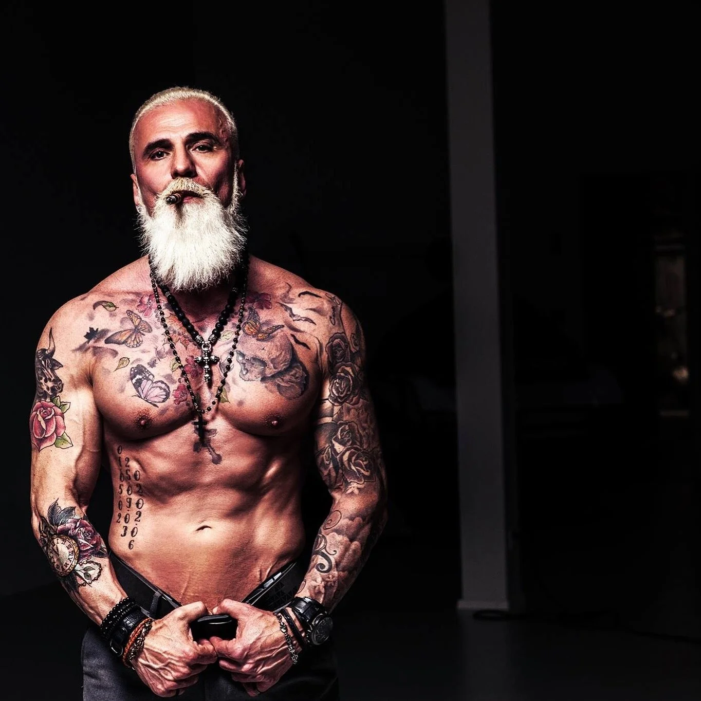 Muscular man with tattoos and a white beard, posing shirtless, wearing necklaces and bracelets, against a dark background.