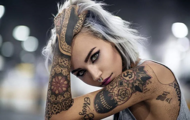 Person with short silver hair and colorful tattoos on arms, wearing a nose ring, with a blurred background.