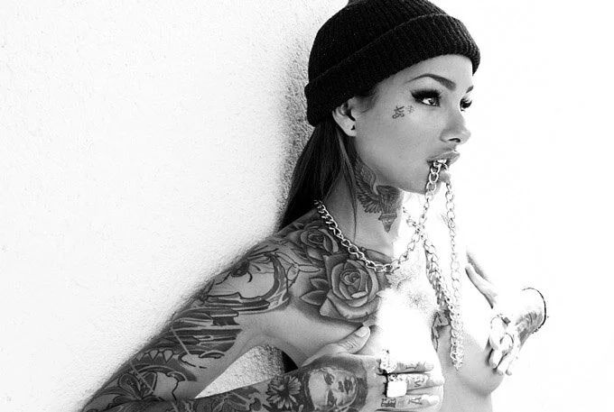Black and white photo of a tattooed woman wearing a beanie, holding a chain in her mouth, and covering her chest with her hands.