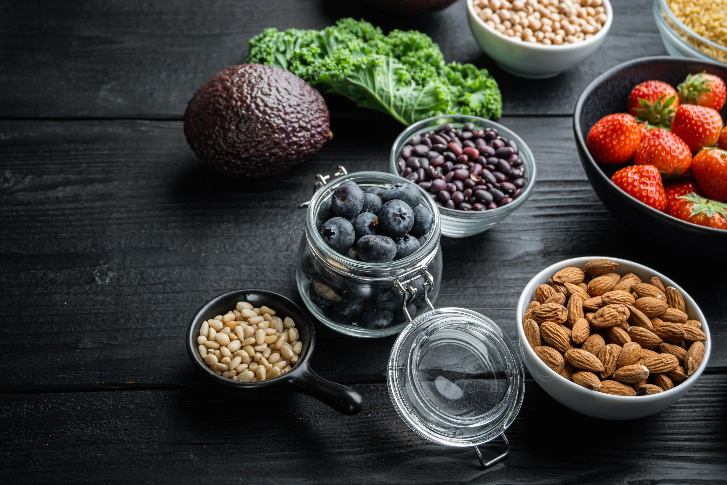 Assorted fresh berries, nuts, seeds, and vegetables on a dark wooden surface, including blueberries, strawberries, black beans, chickpeas, almonds, and an avocado.