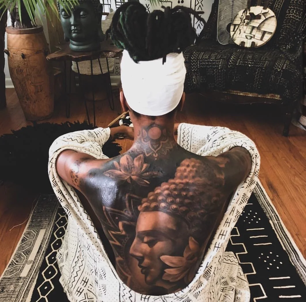 A person with a large Buddha and lotus flower tattoo on their back, sitting on a patterned rug. Their hair is tied up in a white cloth, and they are facing away in a room with tribal decor.