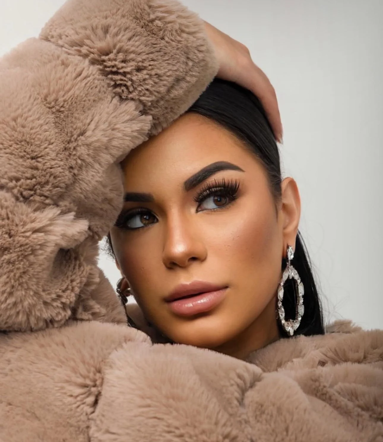 Woman with curly hair wearing a white fluffy robe and a necklace with script pendant, against a brown background.