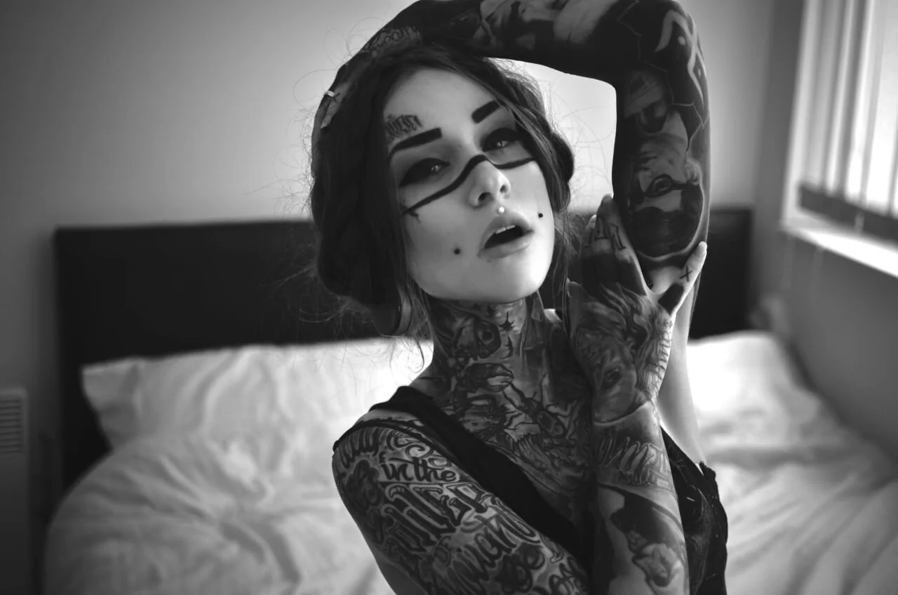 A black and white photo of a person with extensive tattoos, including facial, neck, and arm tattoos, posing with their arm raised, in a bedroom setting.