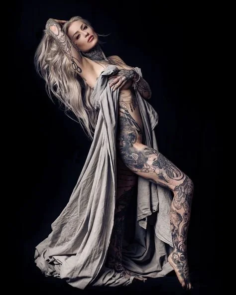 Tattooed person with long hair posing with a draped cloth, against a dark background.