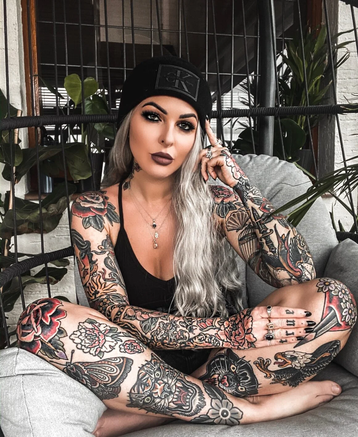 Woman with long silver hair and full-sleeve tattoos sitting cross-legged. She wears a black beanie and black nail polish. Background includes potted plants and a wire mesh.