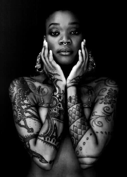 Black and white photo of a person with facial and body tattoos, nose ring, and large earrings, posing with hands near face.
