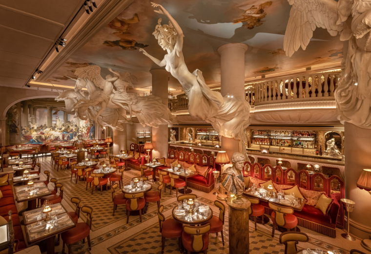 Elegant restaurant interior with large marble sculptures of winged women hanging from the ceiling, plush red seating, and a bar area in the background.