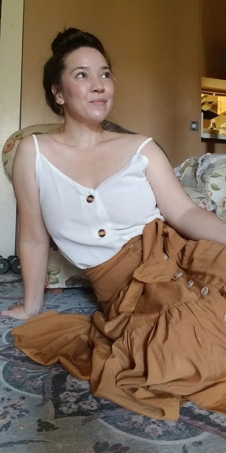 A woman with dark hair styled in a braid, wearing beaded earrings and a black top, smiling at the camera in a home setting.