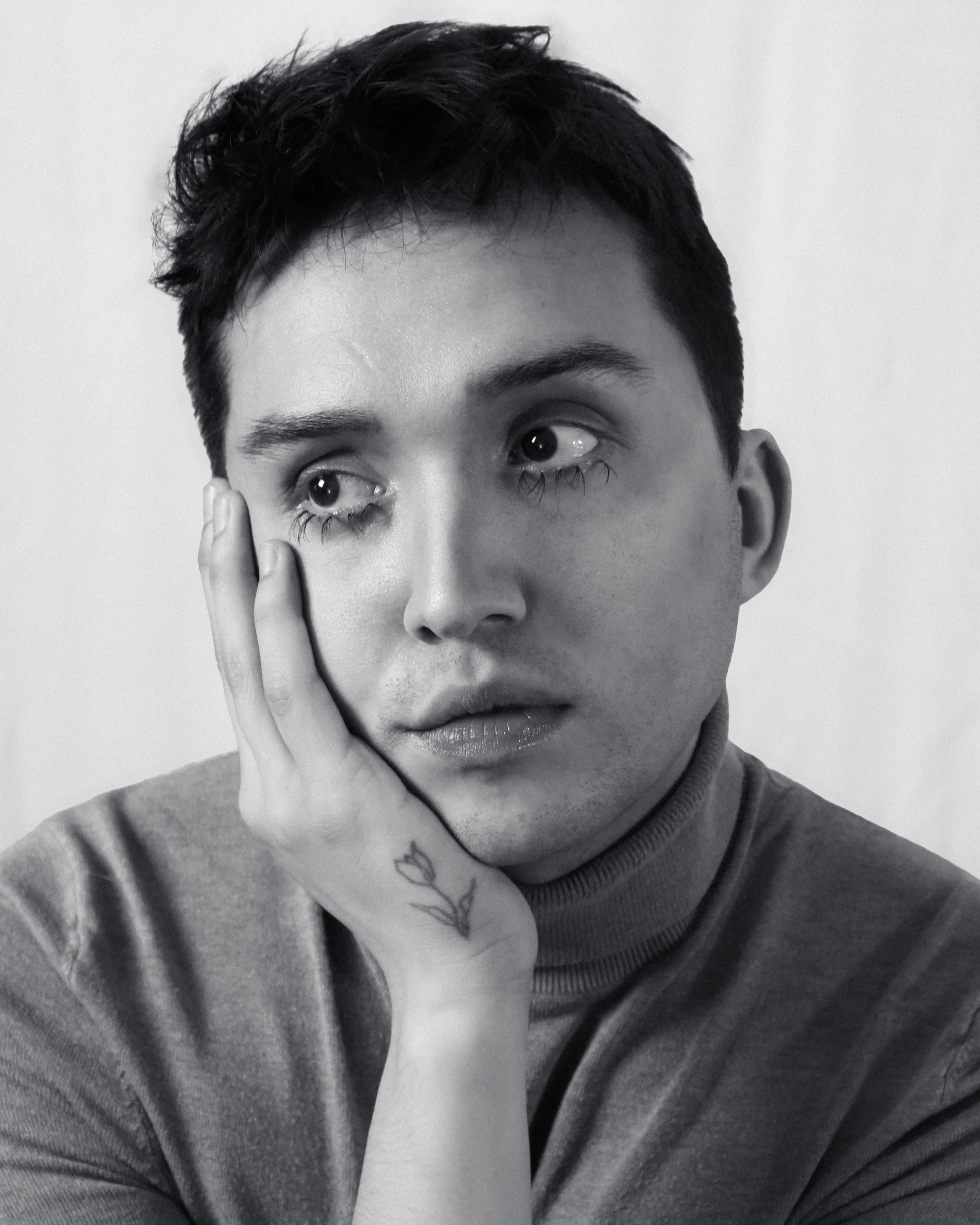 Black-and-white portrait of a young man with short dark hair, resting his face on his hand and looking to the side.