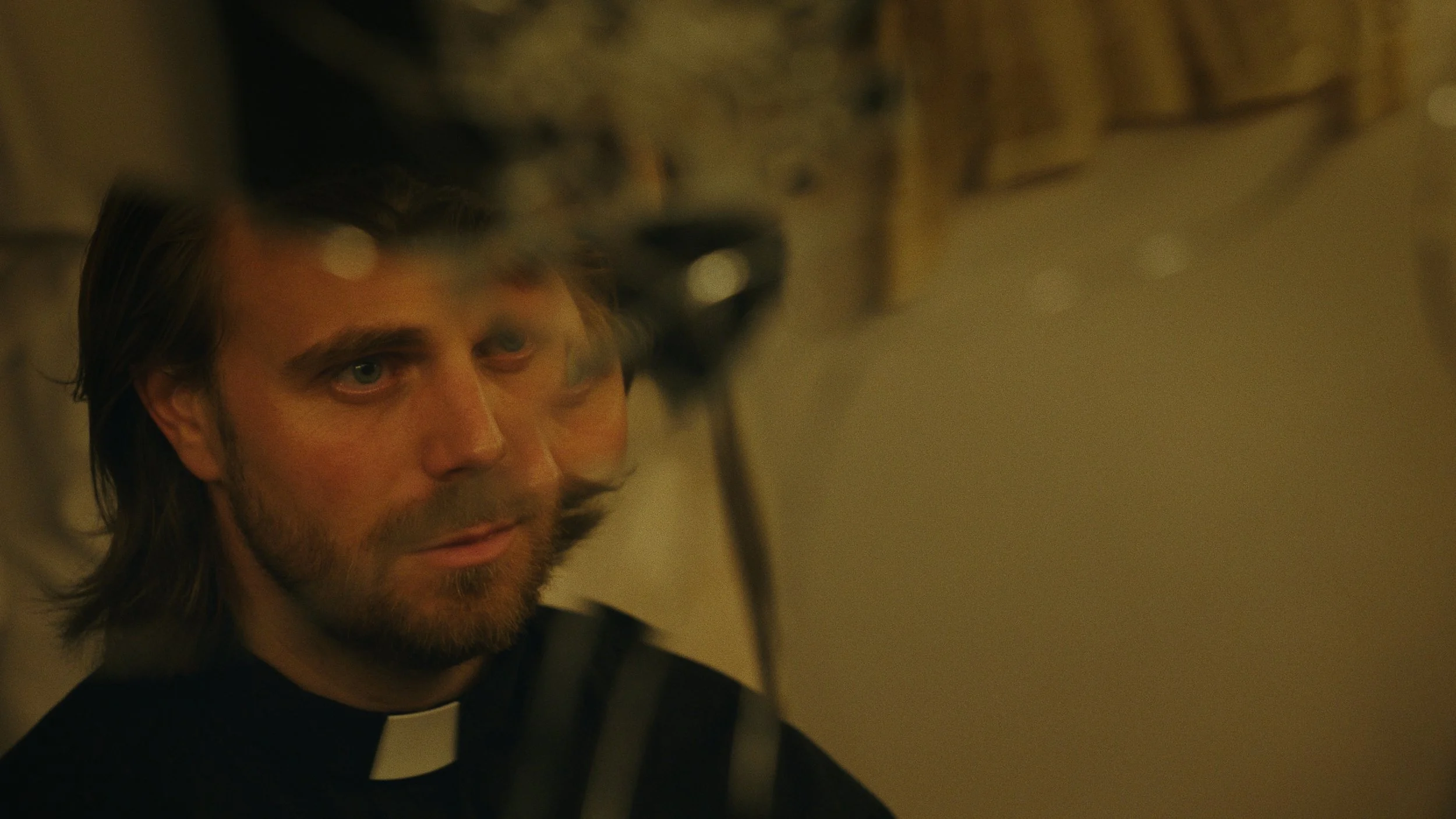 A man with long hair, a beard, and light skin looking to the side, wearing a black shirt and clerical collar, with a blurred background.