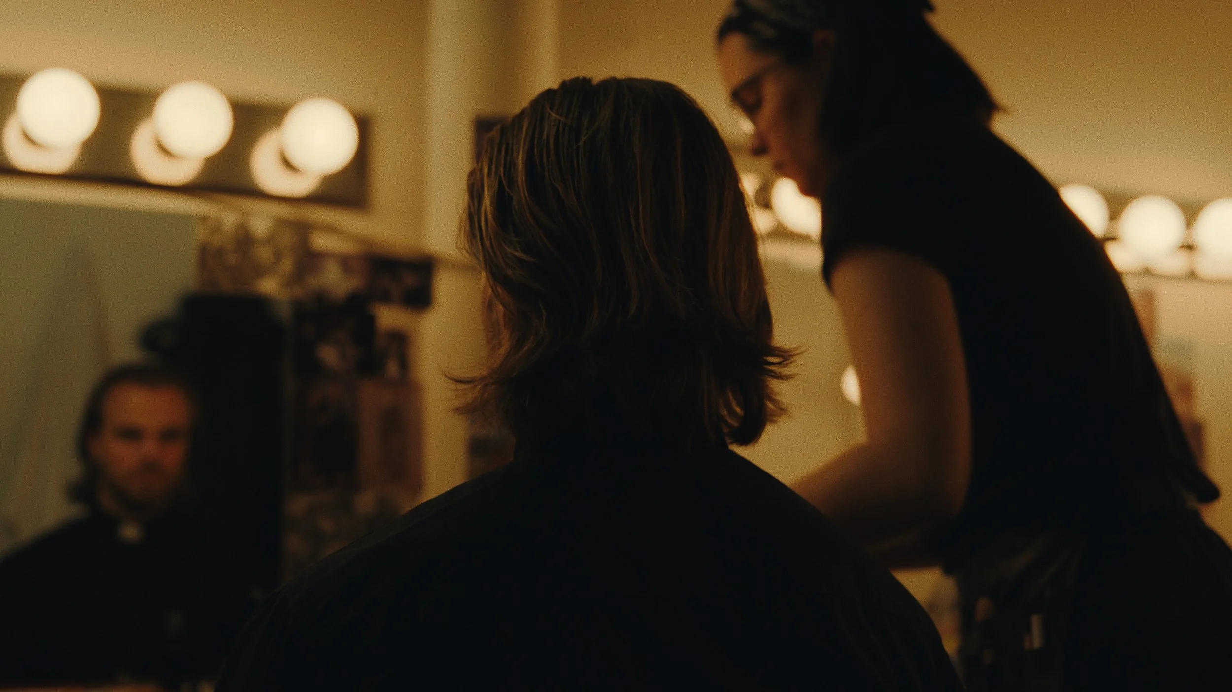 A hairstylist working on a woman's hair in a salon with warm lighting, with another woman and a mirror in the background.