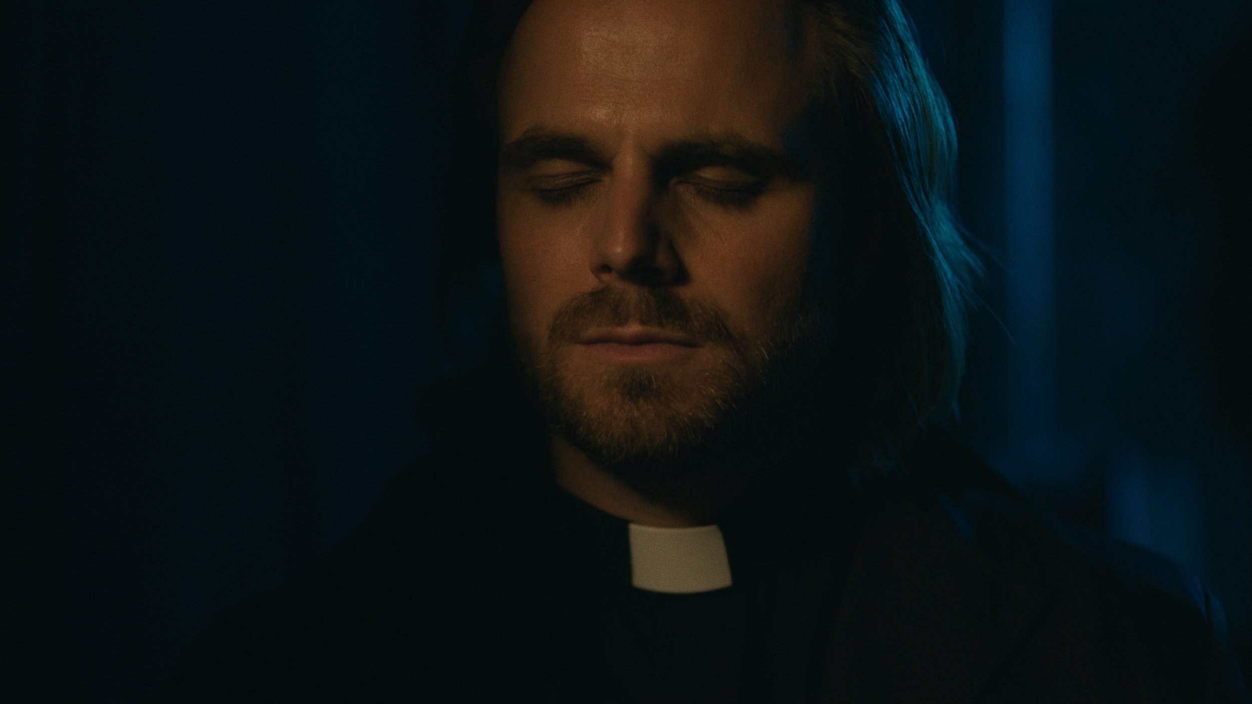 A man with closed eyes, long hair, and a beard dressed as a priest in a dark setting.