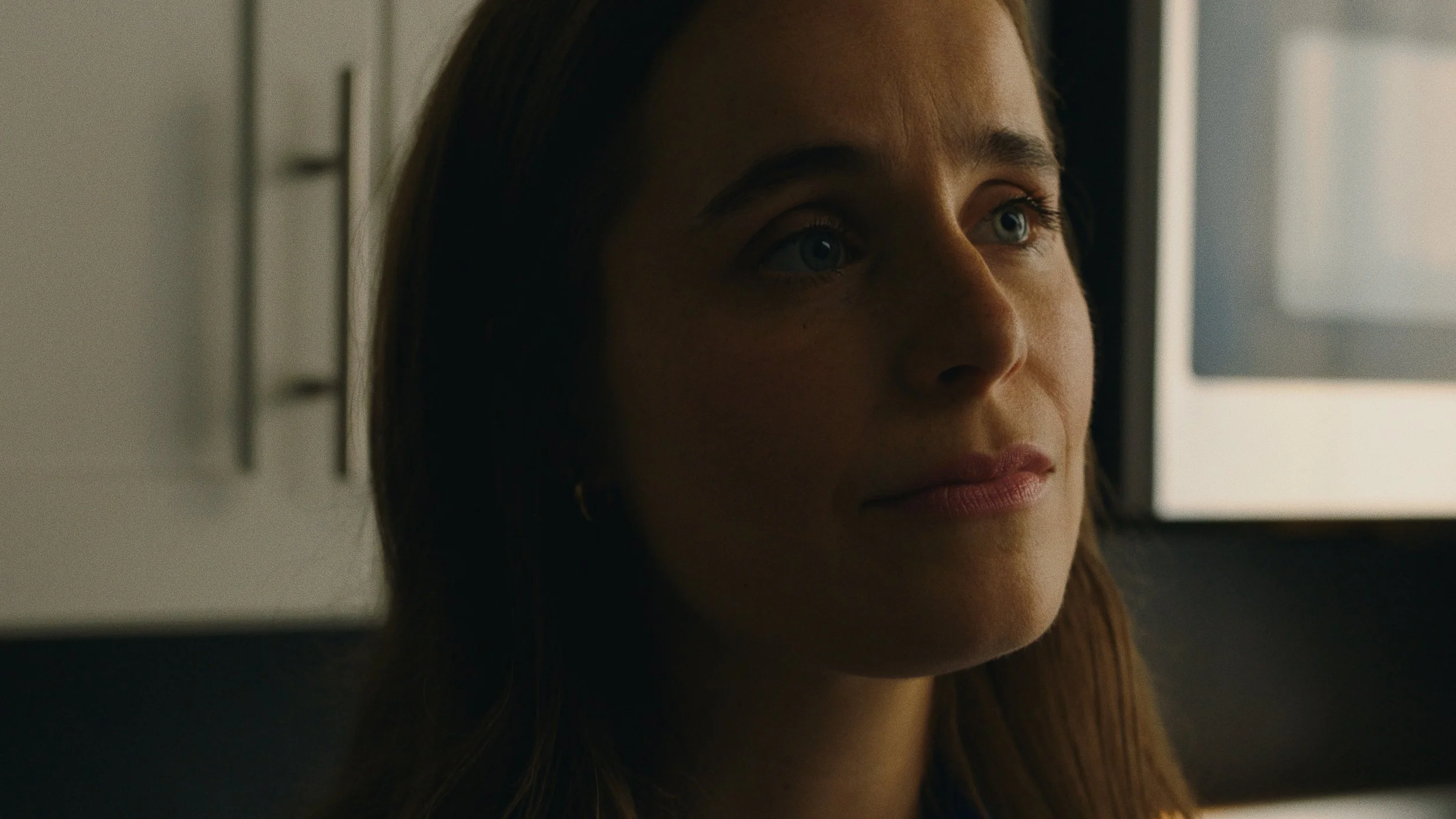 Close-up of a woman with brown hair and blue eyes, looking pensively to the side with soft lighting.