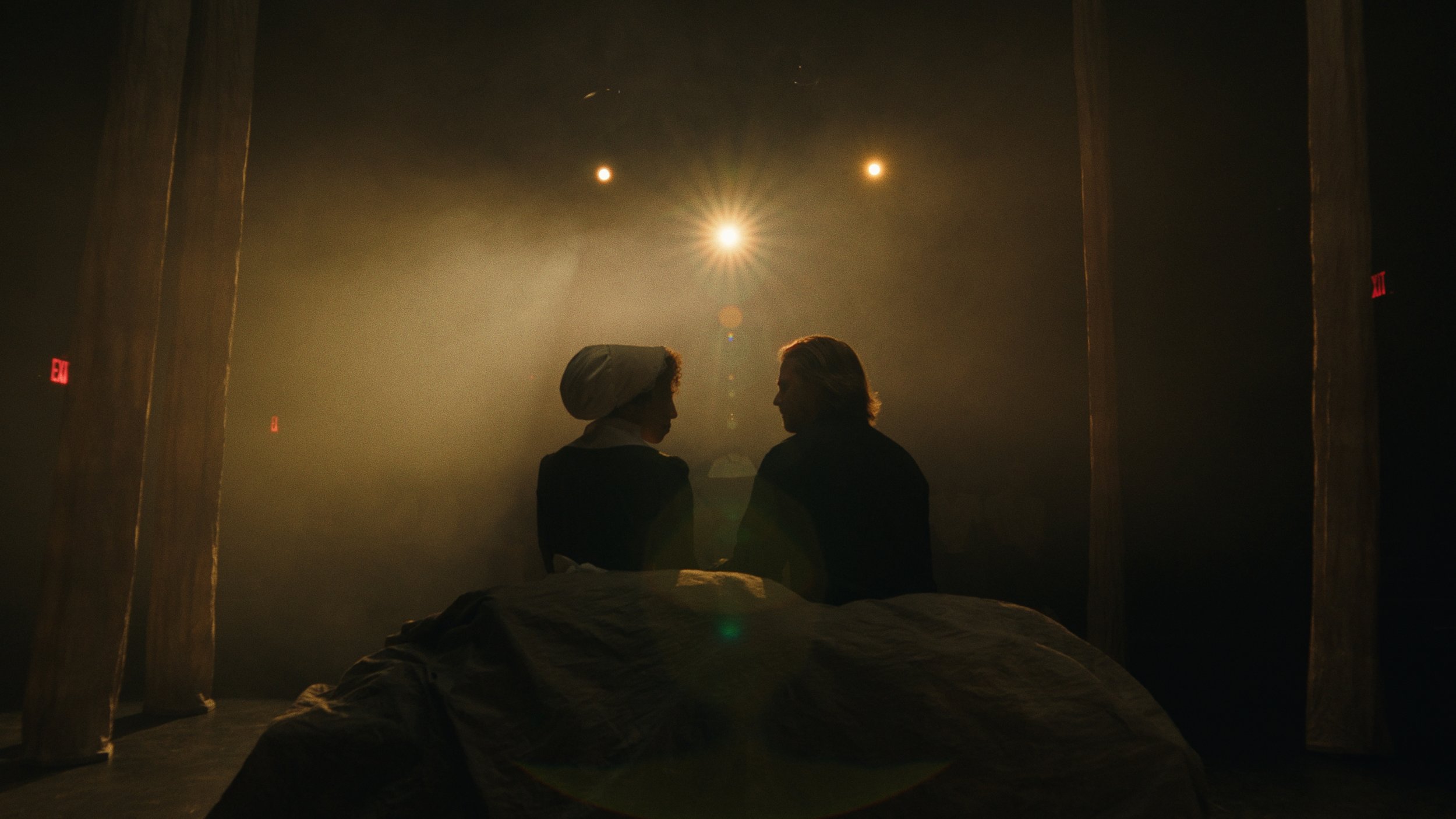 Silhouettes of two people facing each other, sitting on a bed in a dimly lit room with wooden pillars, under a starry sky with three bright celestial objects.