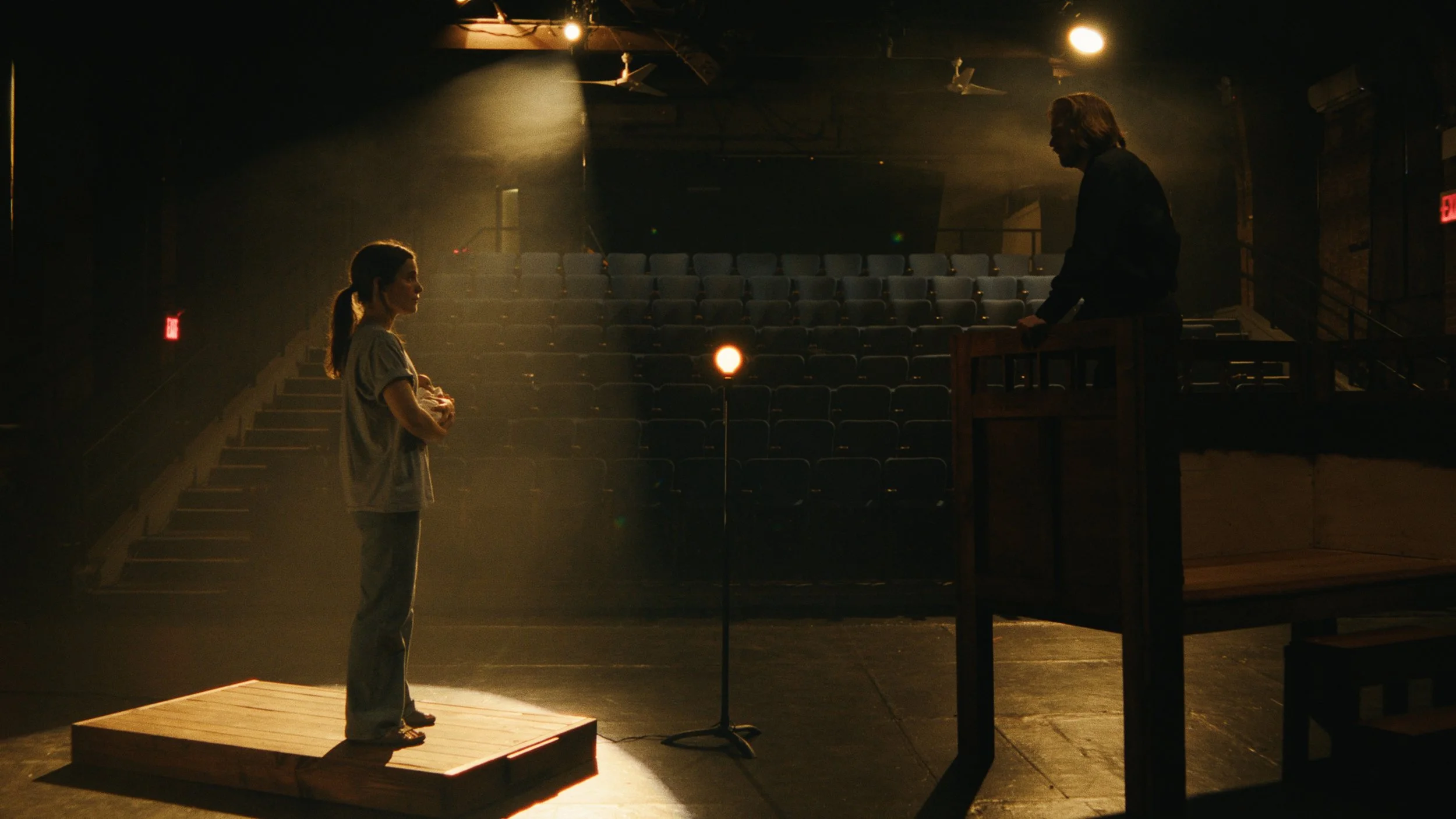 Two actors performing on stage in a theater. The woman stands on a step, looking at the man who is on a raised platform, bathed in warm theatrical lighting.