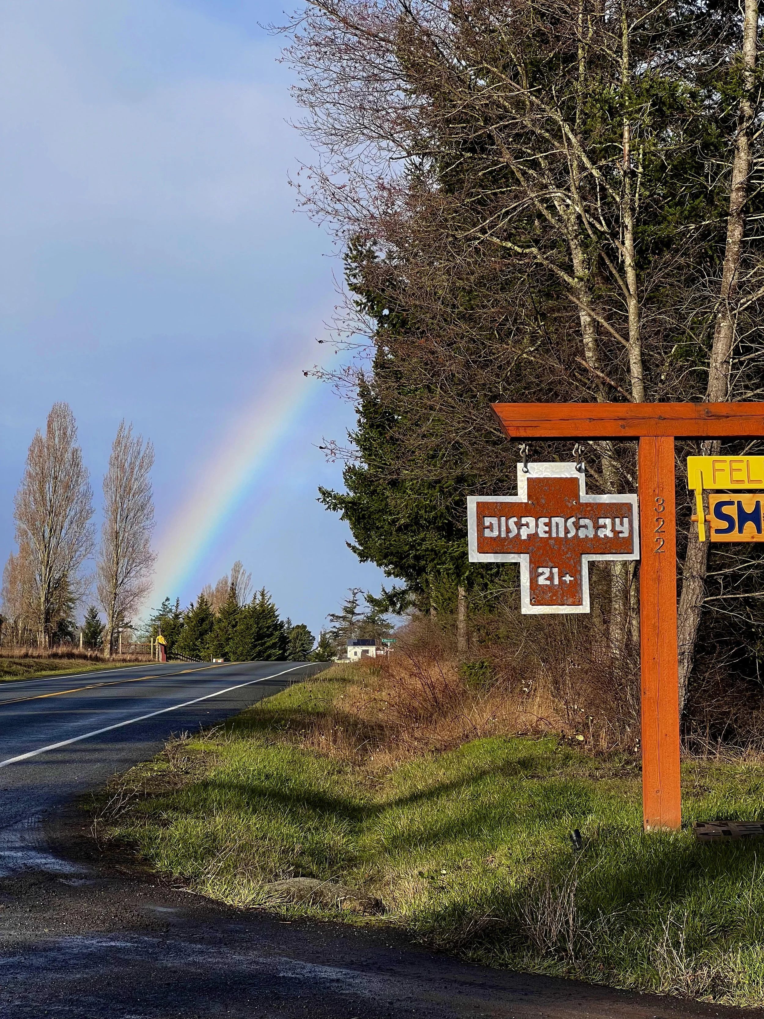 A rural road with a rainbow in the sky, a brown and white roadside sign that reads 'Dispensary 21+', and another sign partially visible behind a wooden post.