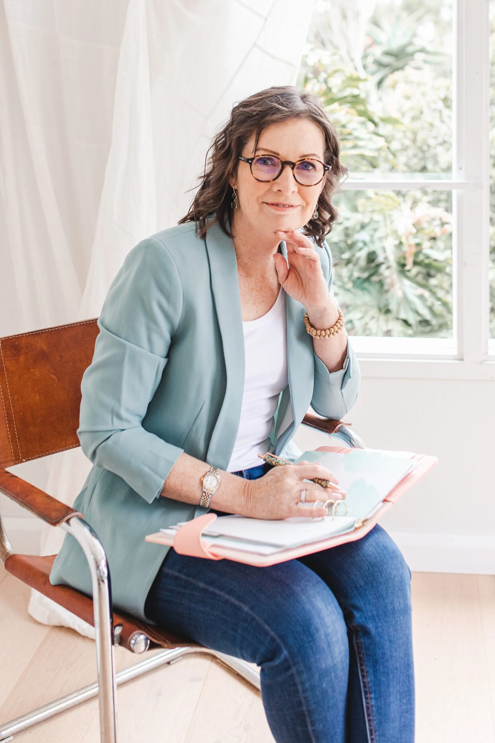 A woman seated on a brown leather chair, wearing glasses, a light blue blazer, and dark jeans. She has a notebook and pen, with a window and greenery in the background.