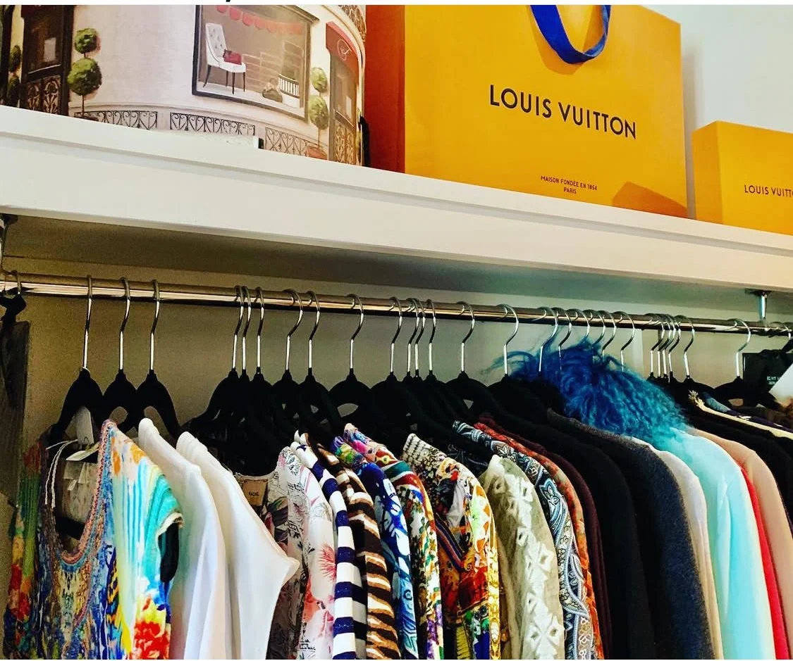 Clothes hanging on a rack in a store with a Louis Vuitton shopping bag on top shelf.