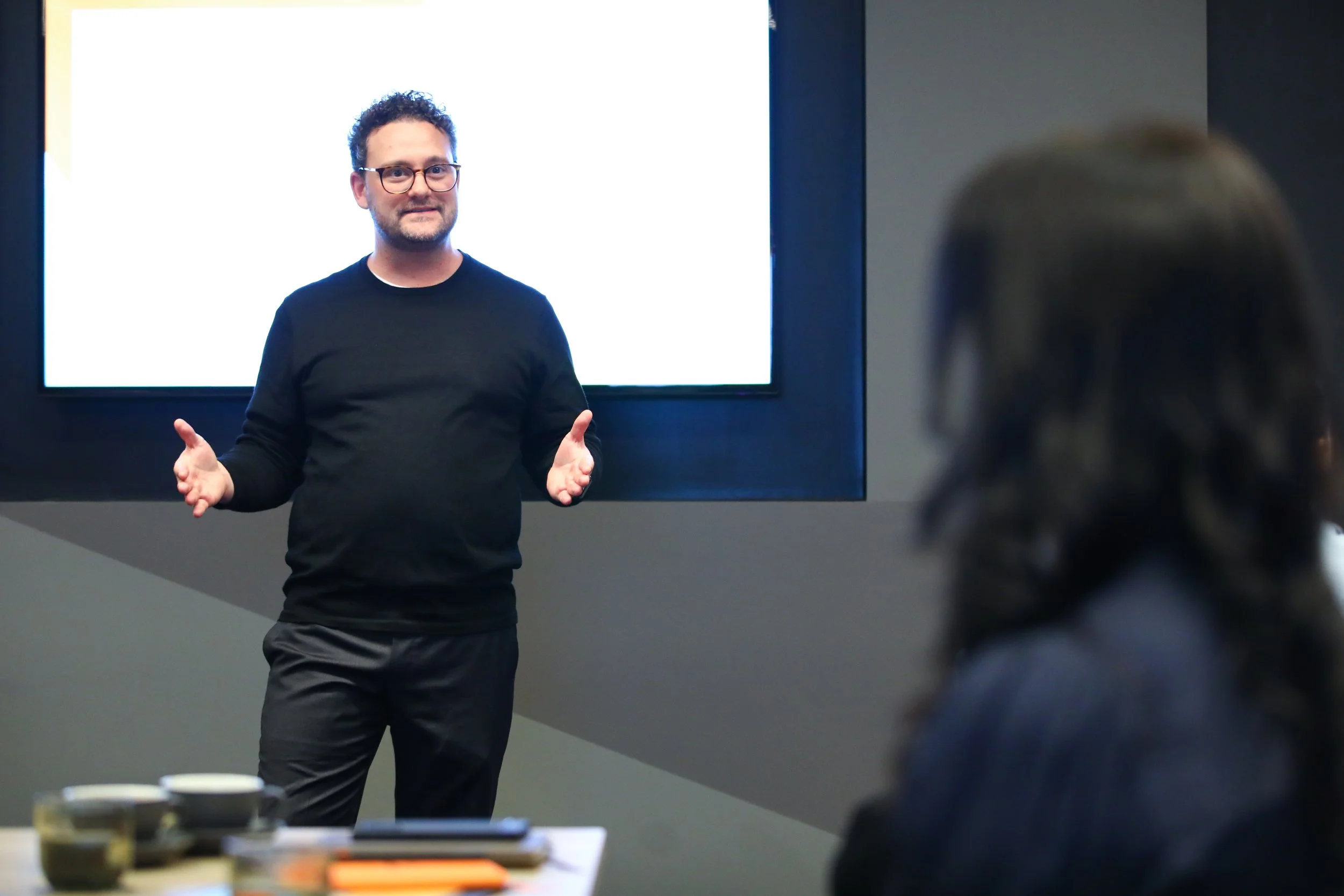 A man with glasses and curly hair wearing a black sweater and black pants giving a presentation in front of a large presentation screen, with an audience member visible in the foreground.