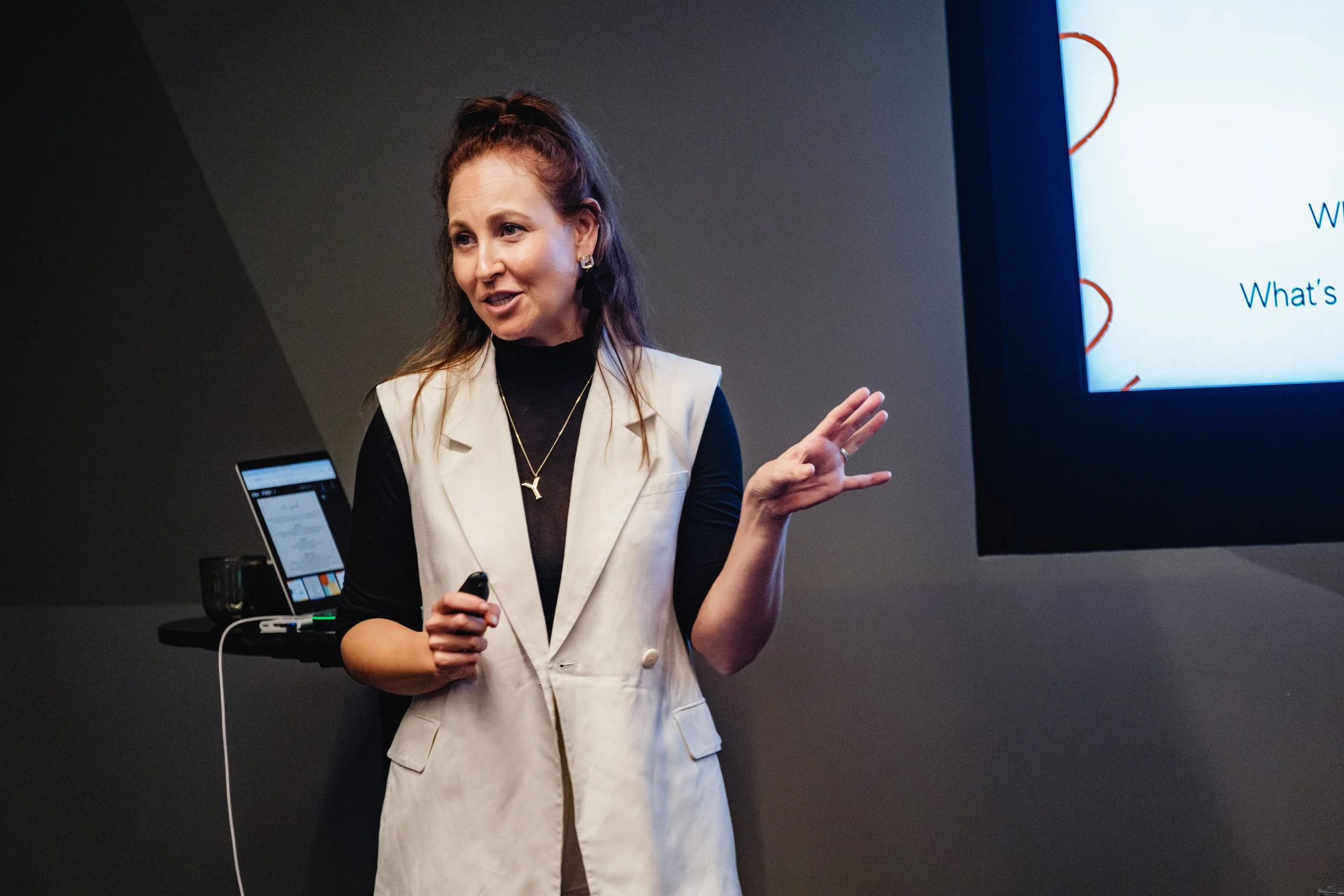 A woman in black attire speaking and gesturing with her hands while seated at a table surrounded by women listening attentively in a conference room.