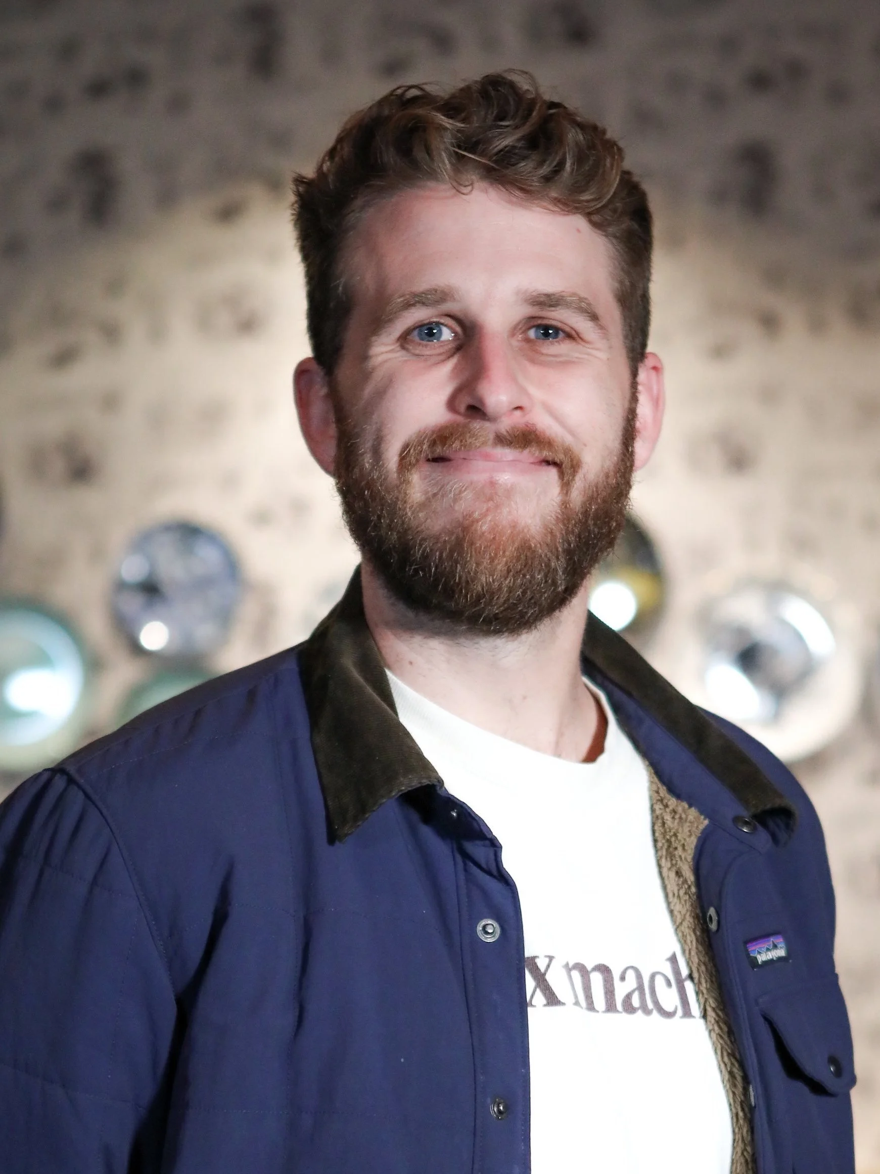 A man with curly hair and a beard smiling, wearing a dark jacket over a white T-shirt with text, standing indoors with a blurred background of lights.