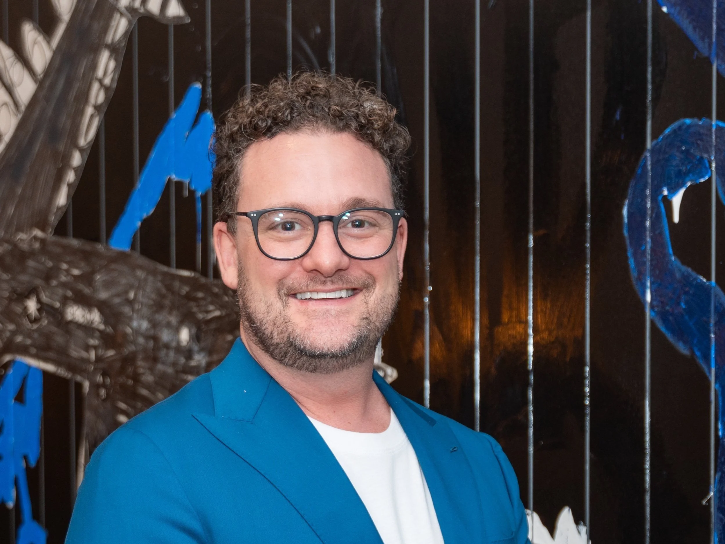 A smiling man with curly hair, glasses, and a beard, wearing a blue blazer over a white shirt, standing in front of an abstract background with blue and black designs.