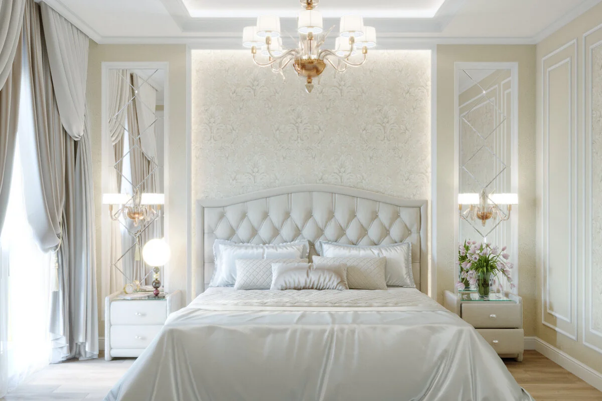 Elegant bedroom with cream-colored walls, white tufted headboard, luxurious bedding, floral arrangement, and chandelier lighting.